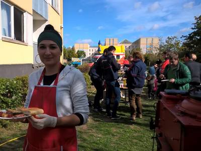 Im Vordergrund steht eine junge Frau lächelnd mit einem Grillteller in der Hand. Im Hintergrund sind Betreuer*innen der Malteser Werke zusehen, die auf einem Gelände vor einer Gemeinschaftsunterkunft stehen.