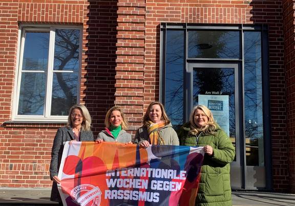 Dezernentin Anja Kerl, Maria Jonas von der Partnerschaft für Demokratie, Madlen Piertza aus der Integrationsarbeit des Landkreises Rostock und Ina-Maria Fahning, Leiterin des Sozialamtes Landkreis Rostock, präsentieren die Fahne vor dem Sitz der Kreisverwaltung in Güstrow. / Landkreis Rostock