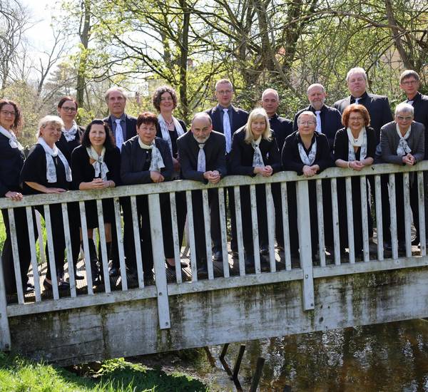 Mitglieder des Chors, gekleidet in schwarze Jacken mit weissen Schals, stehen auf einer kleinen weissen Brücke und sehen in das Objektiv