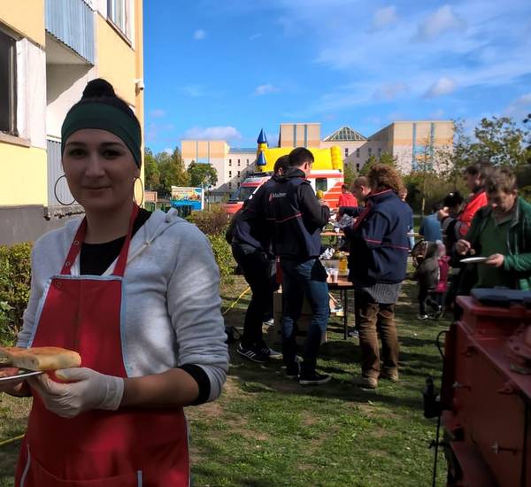 Im Vordergrund steht eine junge Frau lächelnd mit einem Grillteller in der Hand. Im Hintergrund sind Betreuer*innen der Malteser Werke zusehen, die auf einem Gelände vor einer Gemeinschaftsunterkunft stehen.